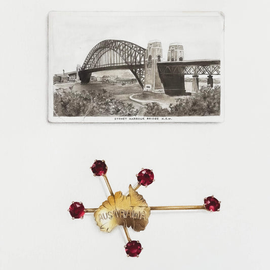 Gold brooch with red gemstones and 'Australia' text on a white background with vintage postcard of the Sydney Harbour Bridge