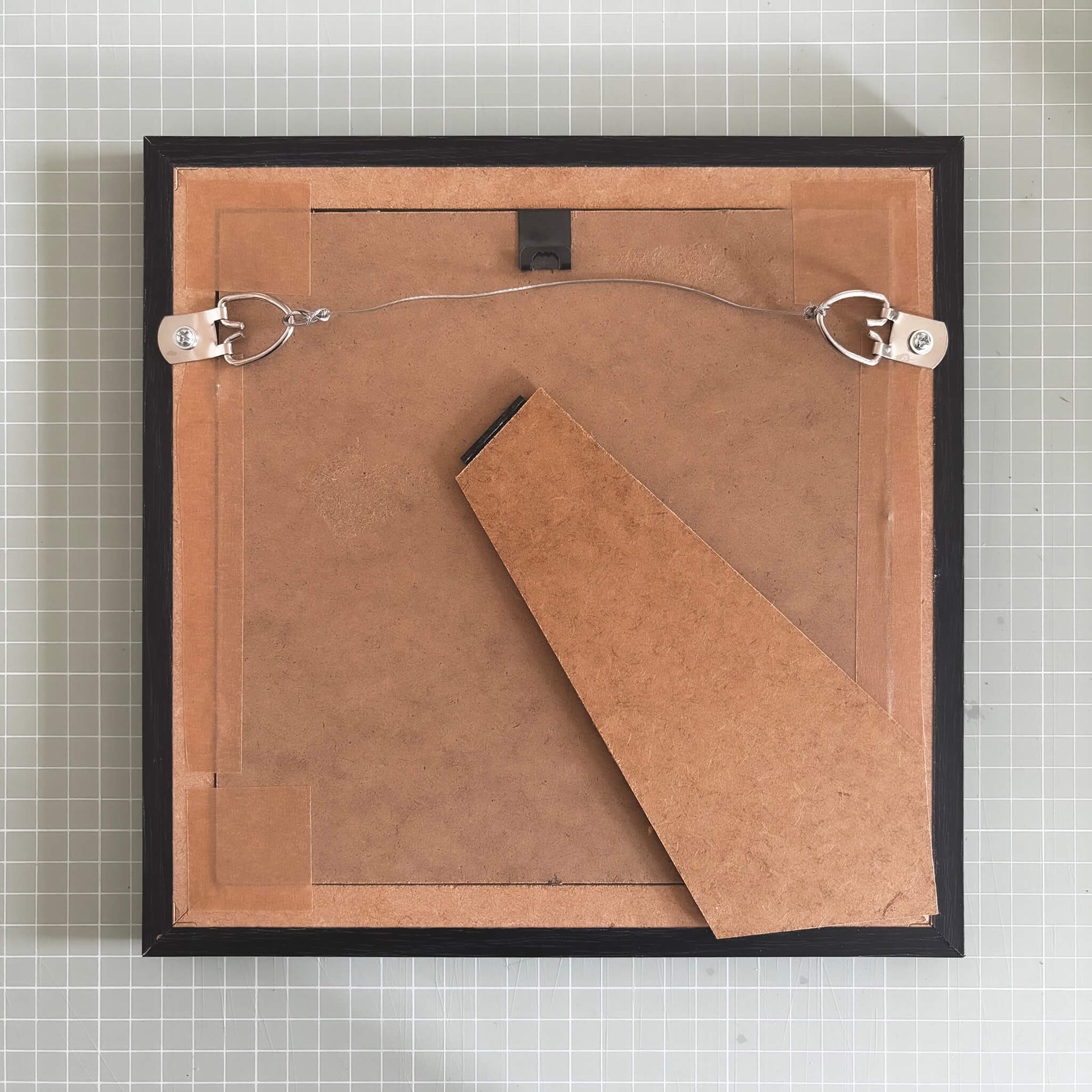 Backside of a framed artwork with cardboard and wire, on a tiled wall background.