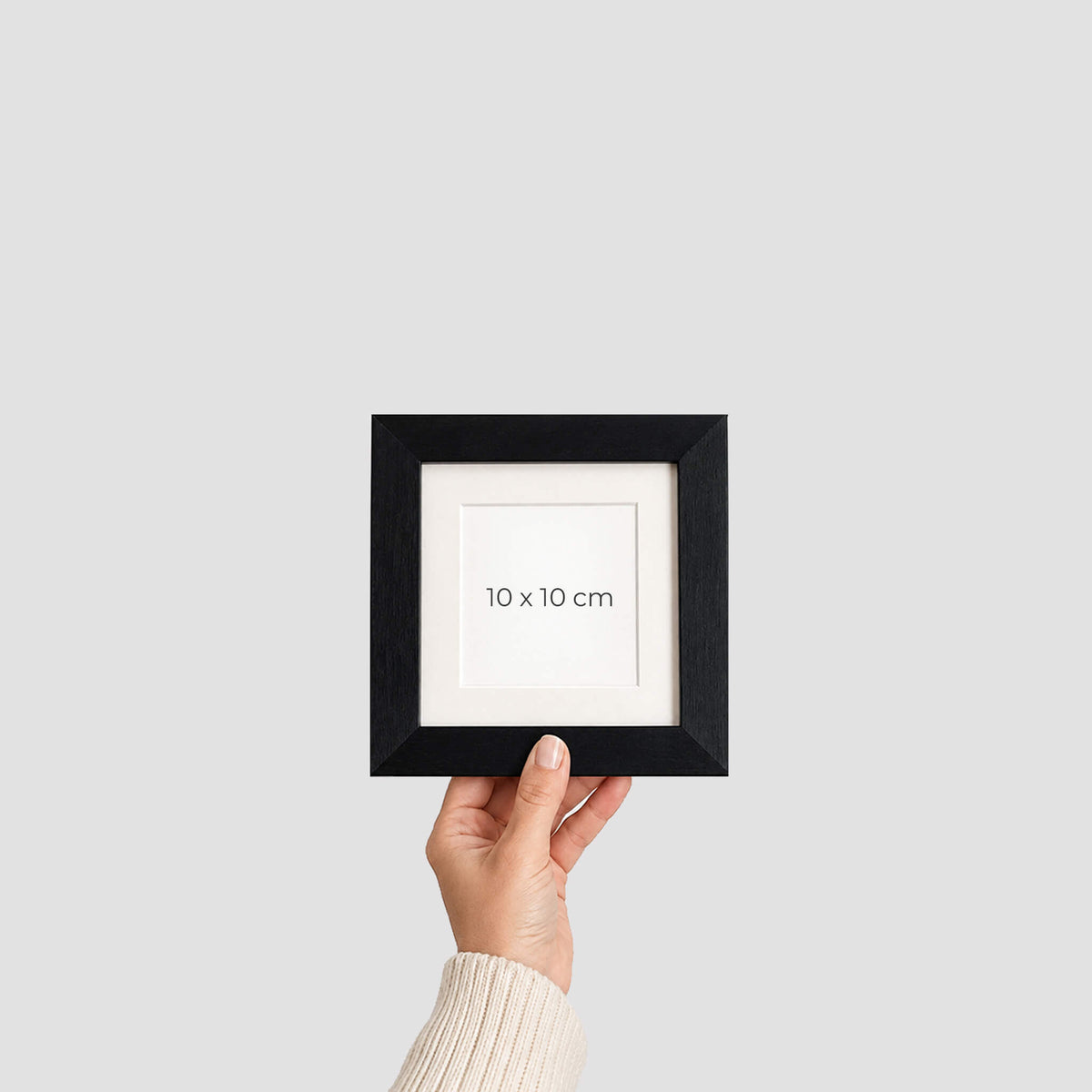 A hand holding a small black picture frame with a white mat and the text '10 x 10 cm' printed in the center, against a light grey background.