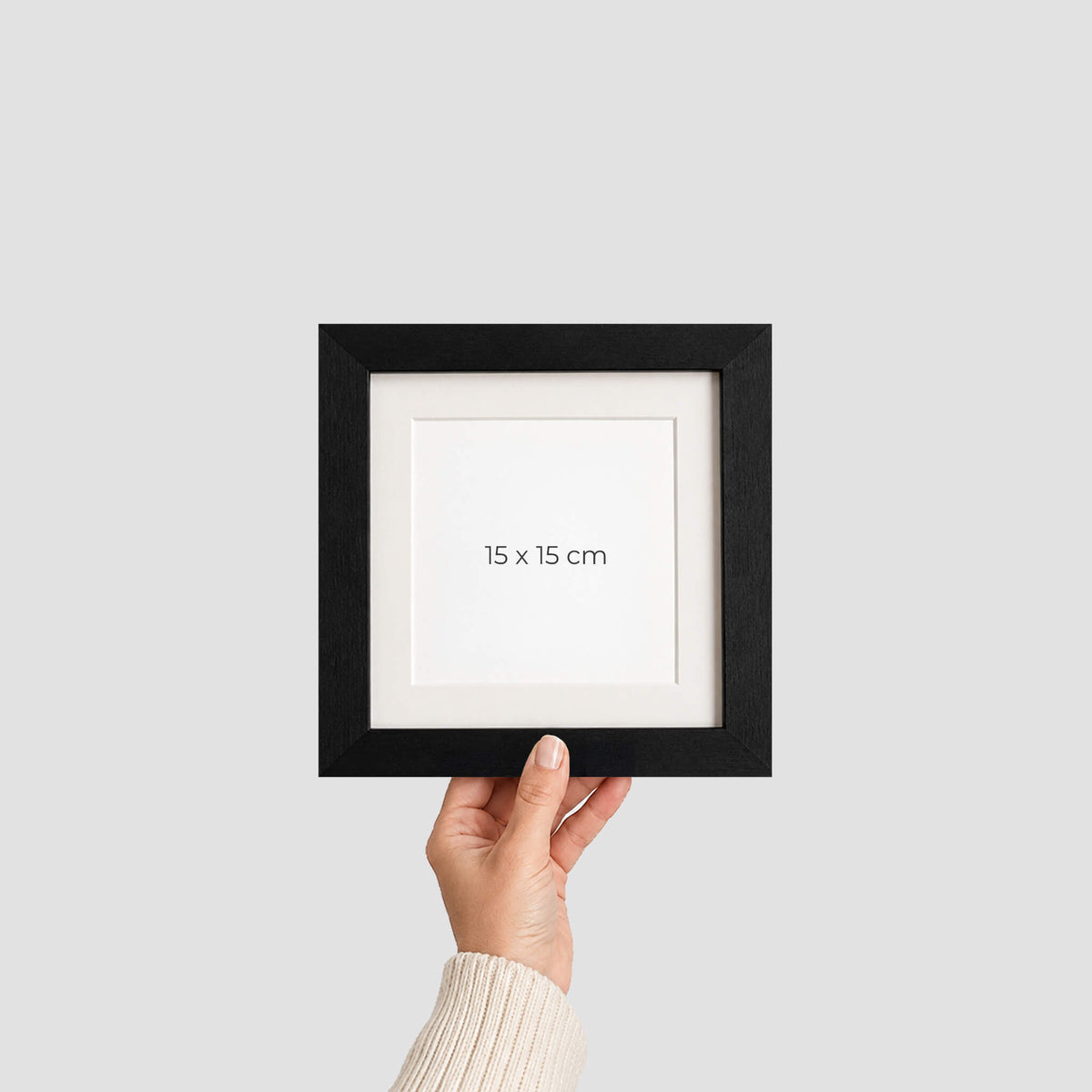 A hand holding a small black picture frame with a white mat and the text '15 x 15 cm' printed in the centre, against a light grey background.