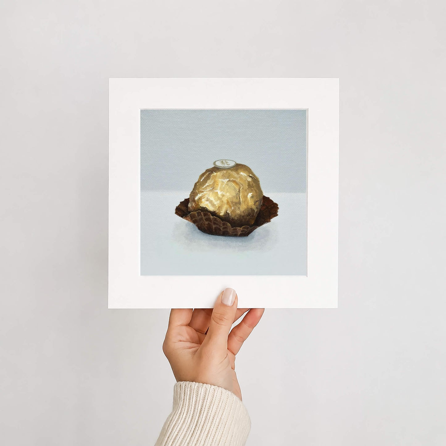 Hand holding a framed picture of a gold foiled chocolate on a light gray background
