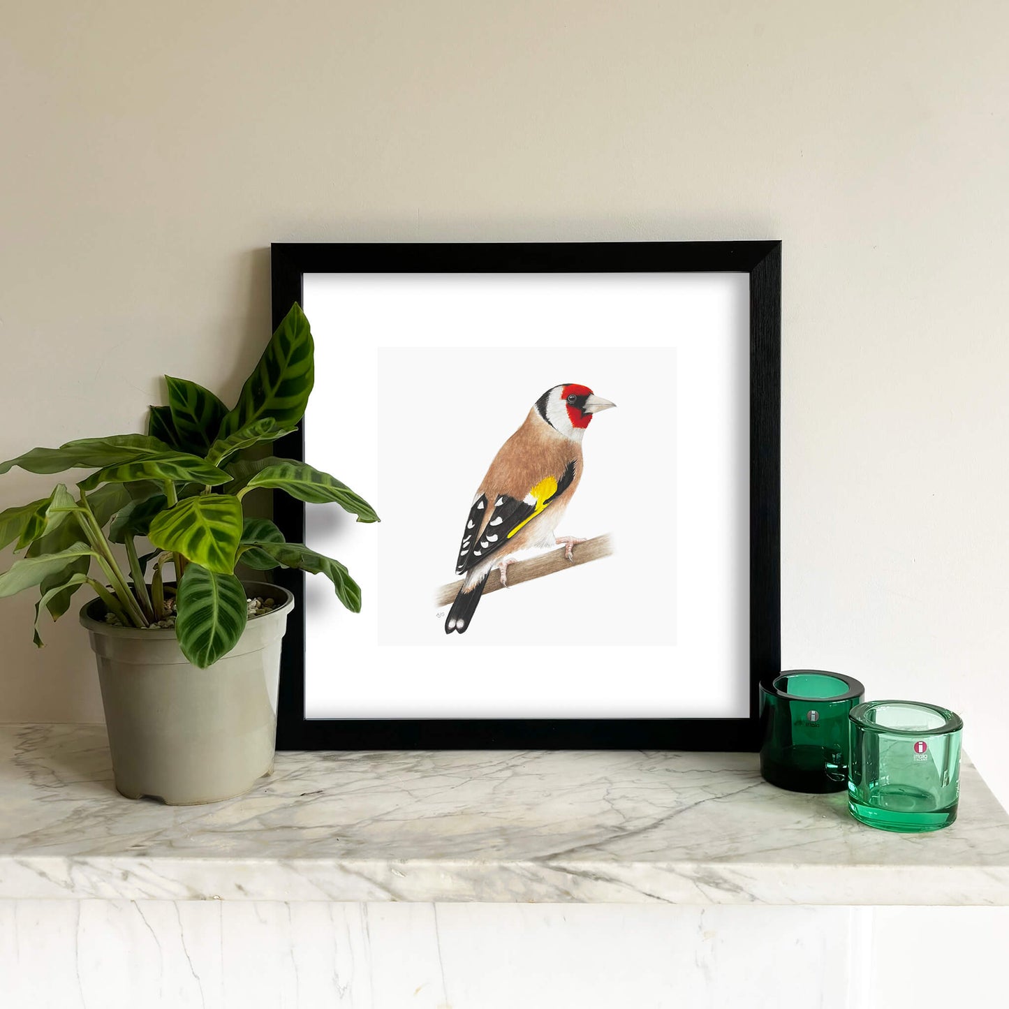 Framed print of a goldfinch bird on a branch with a plant and glasses on a mantlepiece.