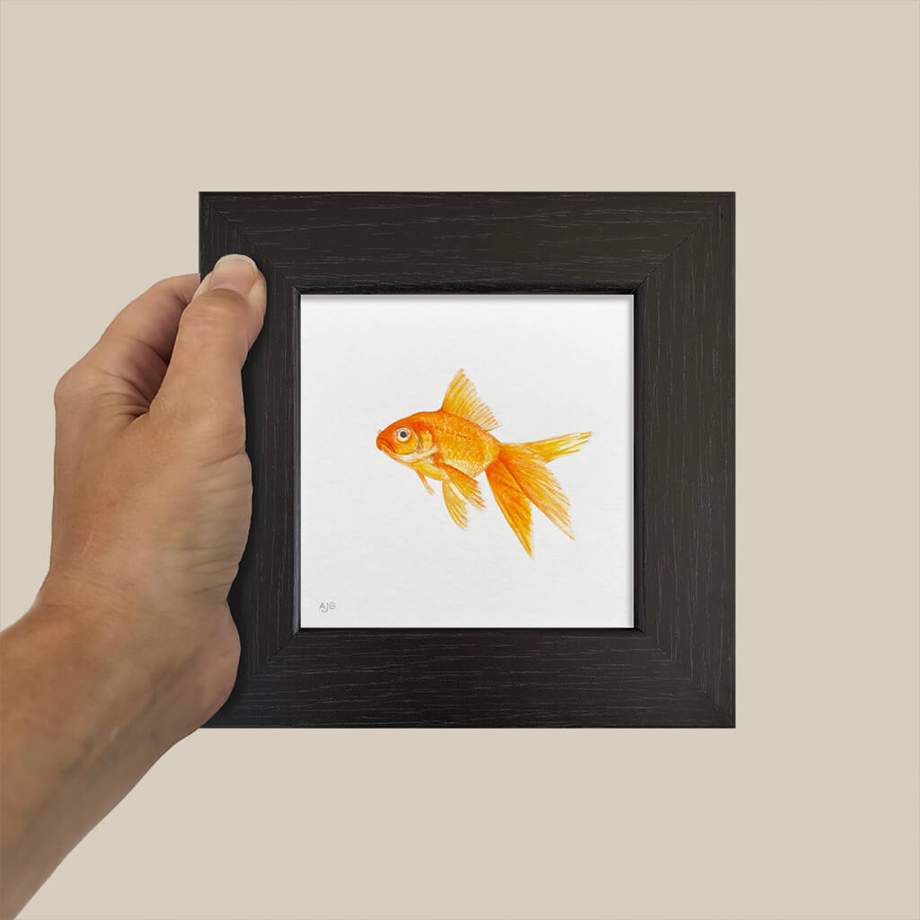 Hand holding a small framed painting of a goldfish against a beige background