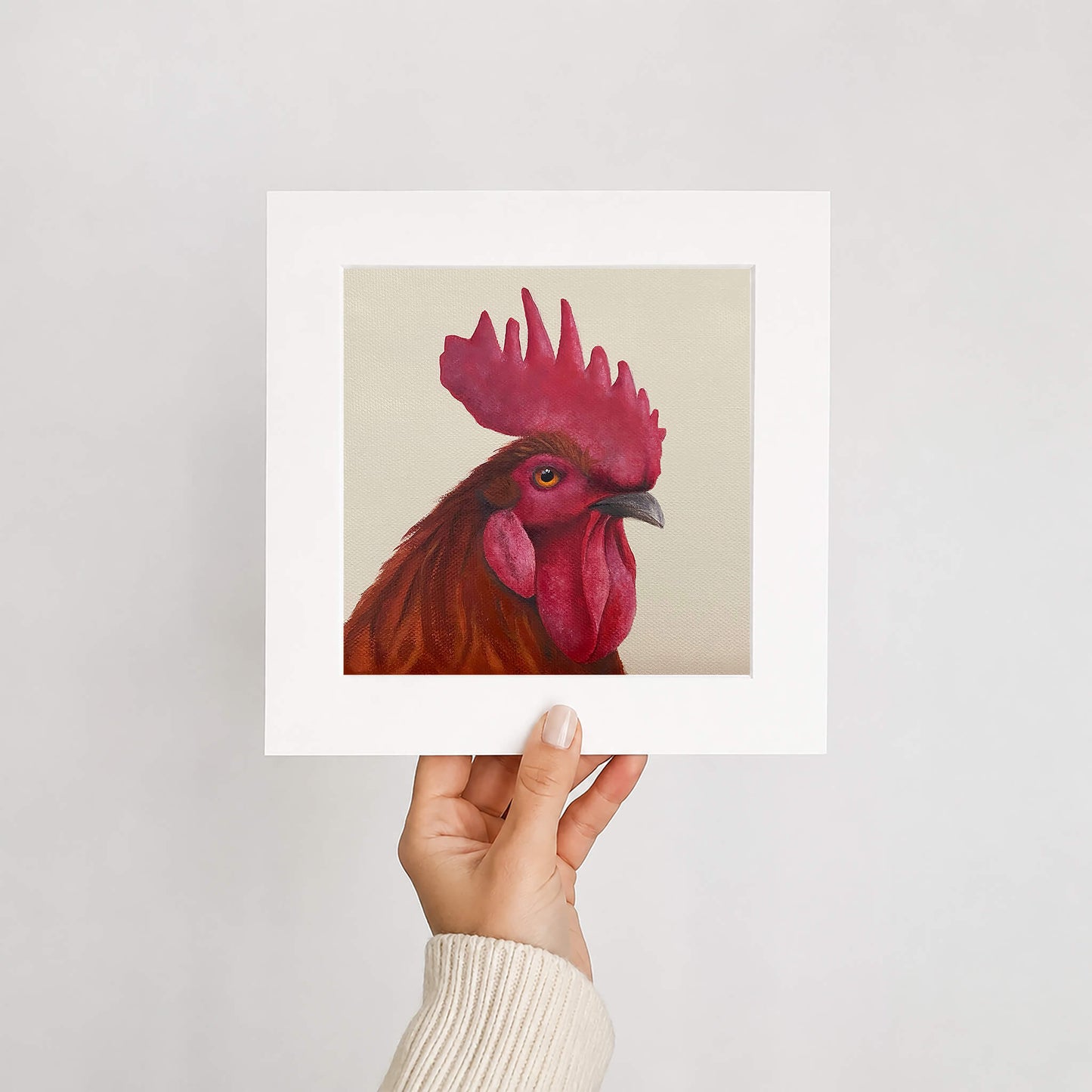 Hand holding a print of a rooster with a red comb against a light gray background