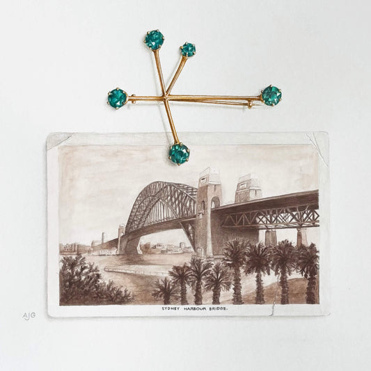 An original gouache painting of a gold brooch with teal green gemstones on a vintage Sydney Harbour Bridge postcard