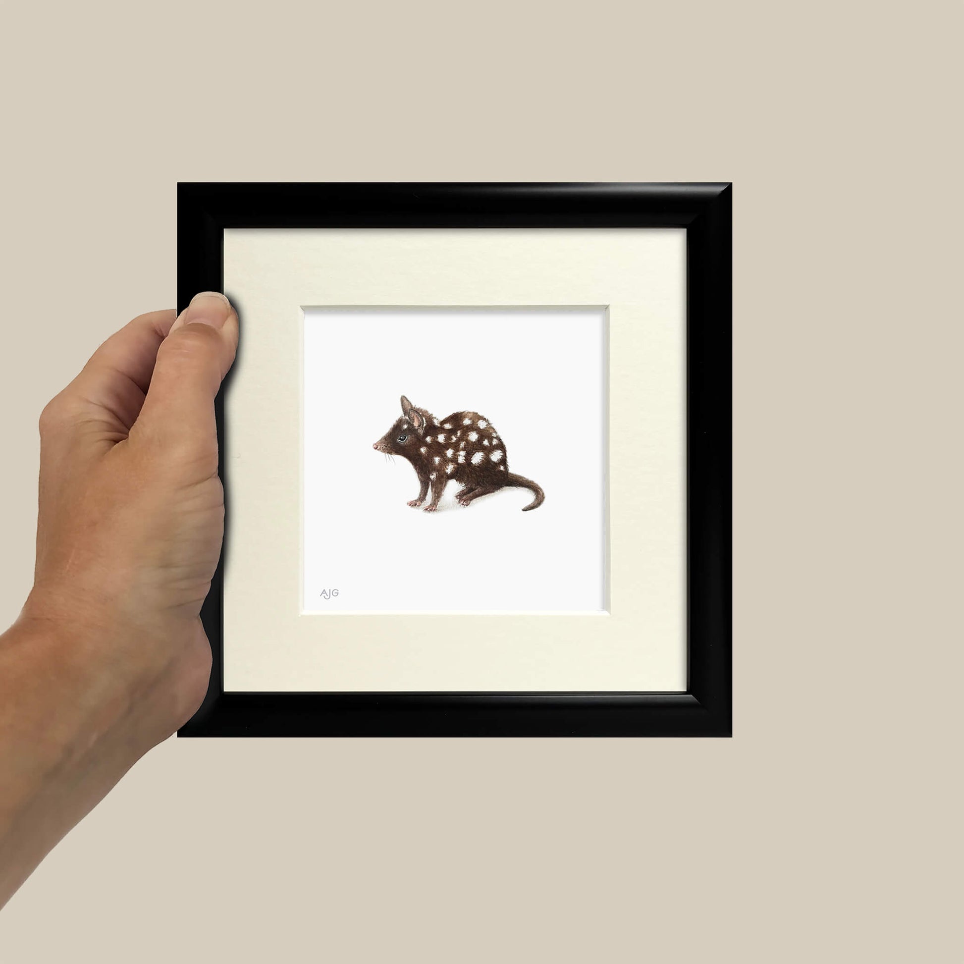 Hand holding a framed picture of a spotted animal illustration on a beige wall.