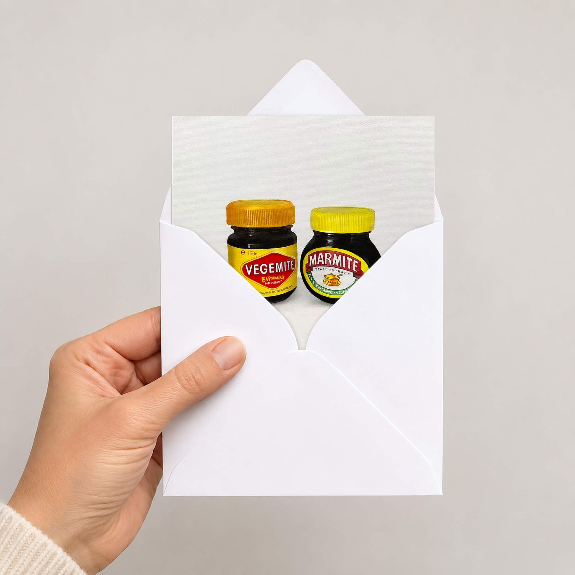 A hand holds a white envelope against a light grey background. Inside the envelope is a printed card featuring two small jars of spread: a Vegemite jar with a yellow lid and a Marmite jar with a yellow lid.