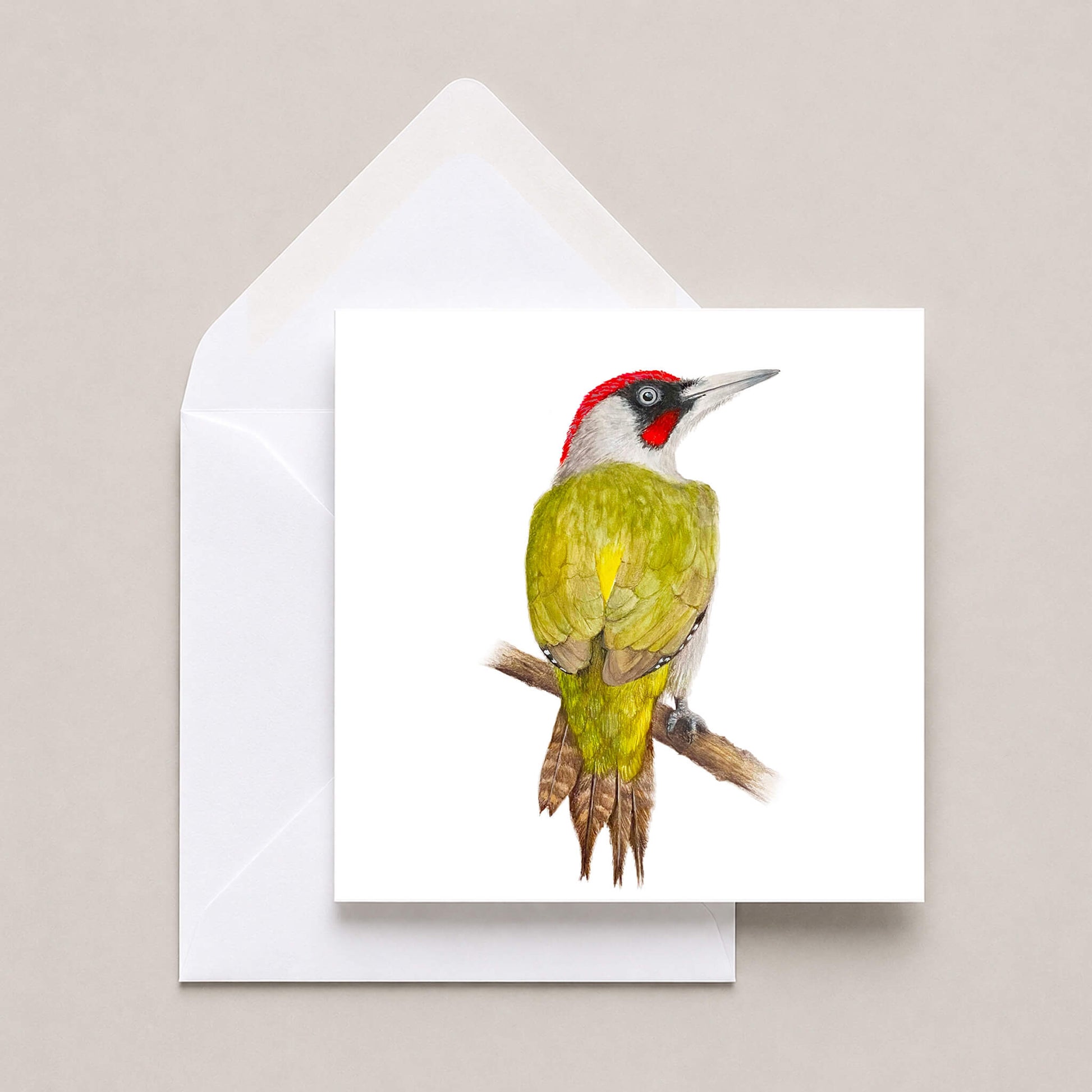 A square white greeting card featuring a painting of a green woodpecker with a red crown, shown with a white envelope on a light beige background.