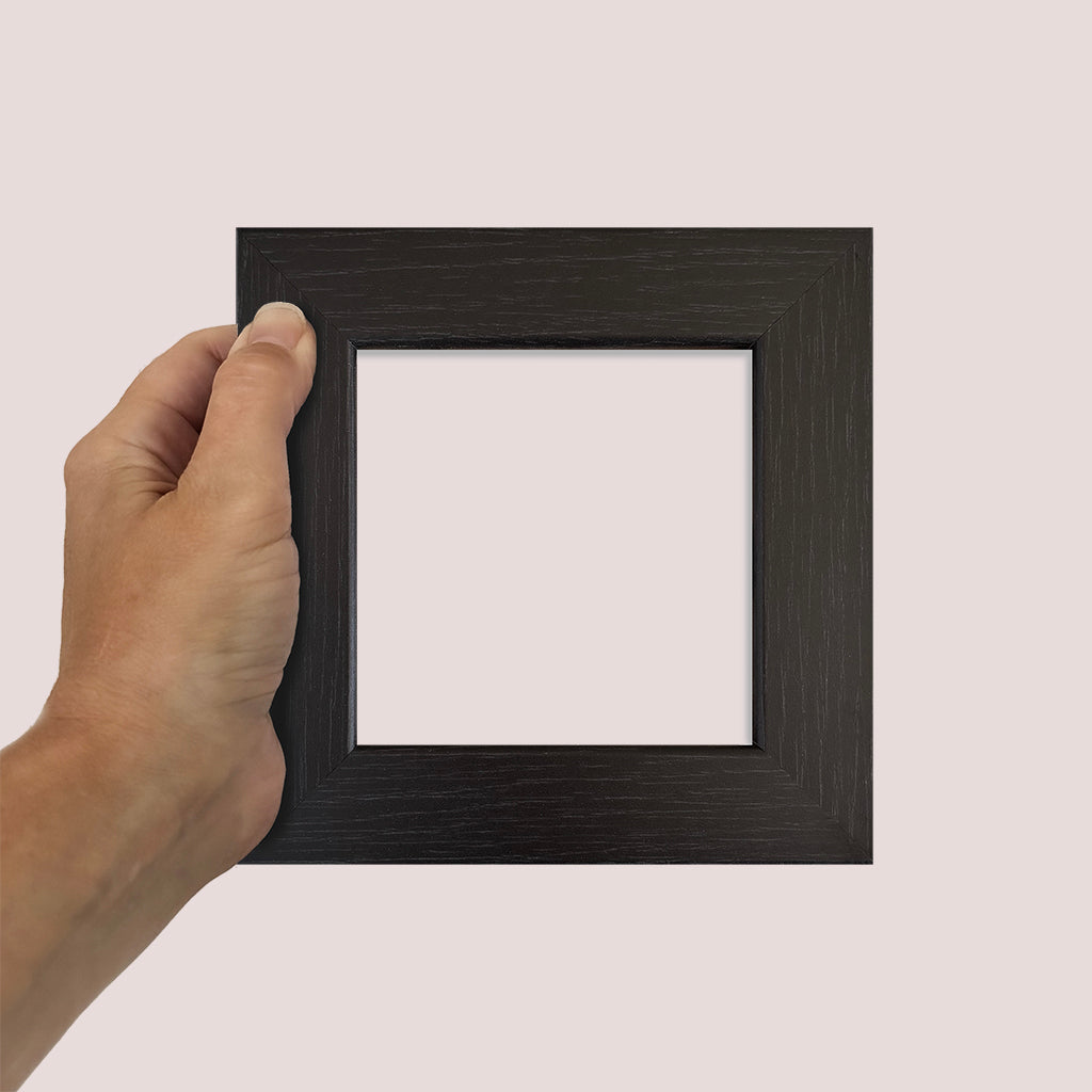 Hand holding a small square black picture frame against a plain background