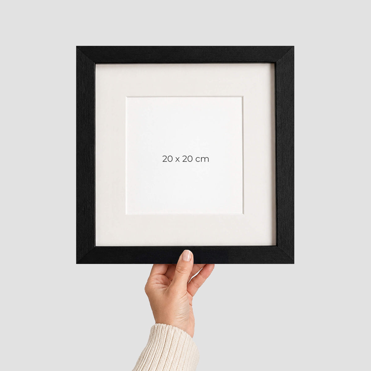 A hand holding a black picture frame with a white mat and the text '20 x 20 cm' printed in the centre, against a light grey background.