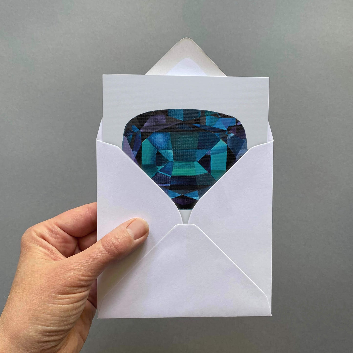 Hand holding a white envelope with an alexandrite gemstone inside against a grey background