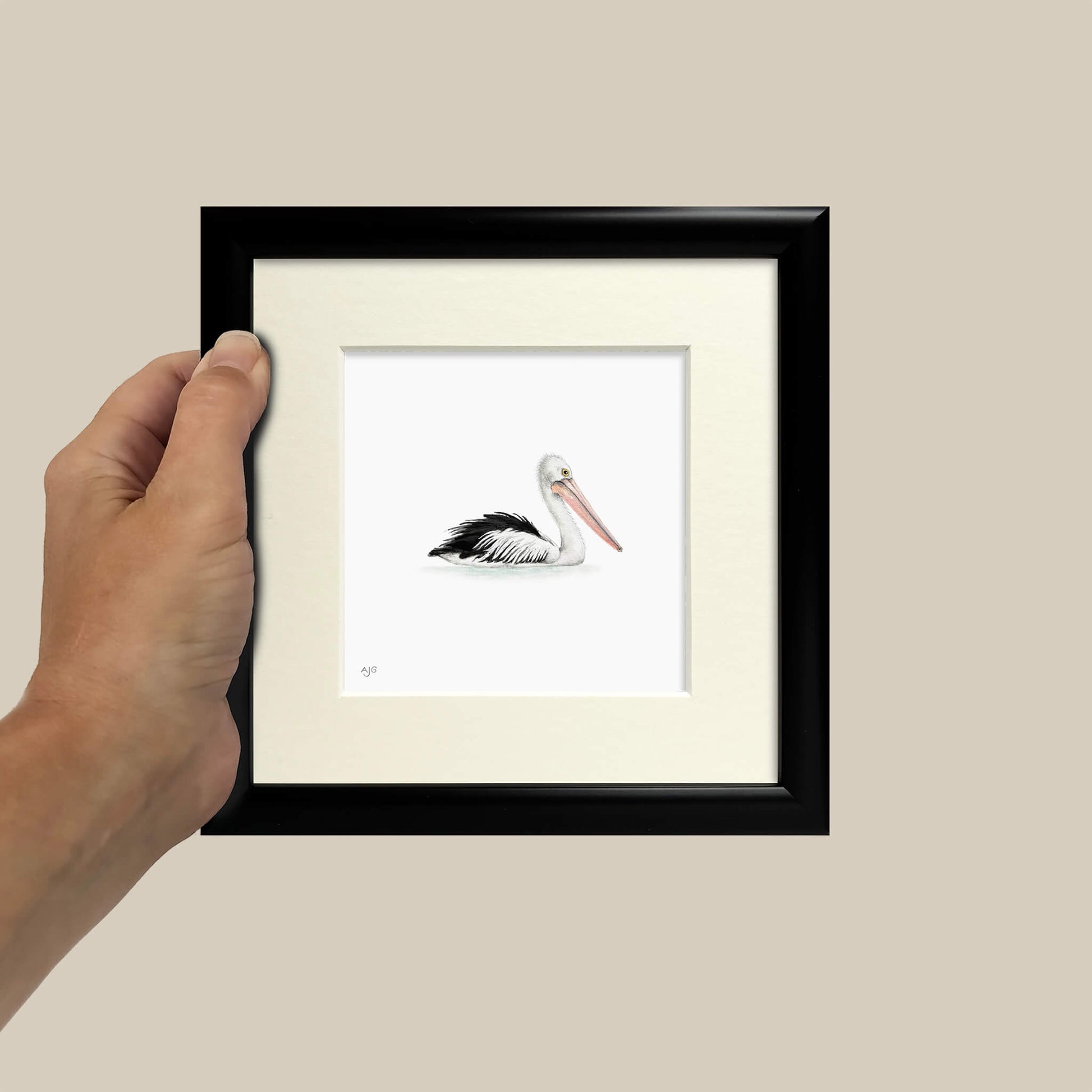 Framed artwork of a pelican held by a hand against a beige background