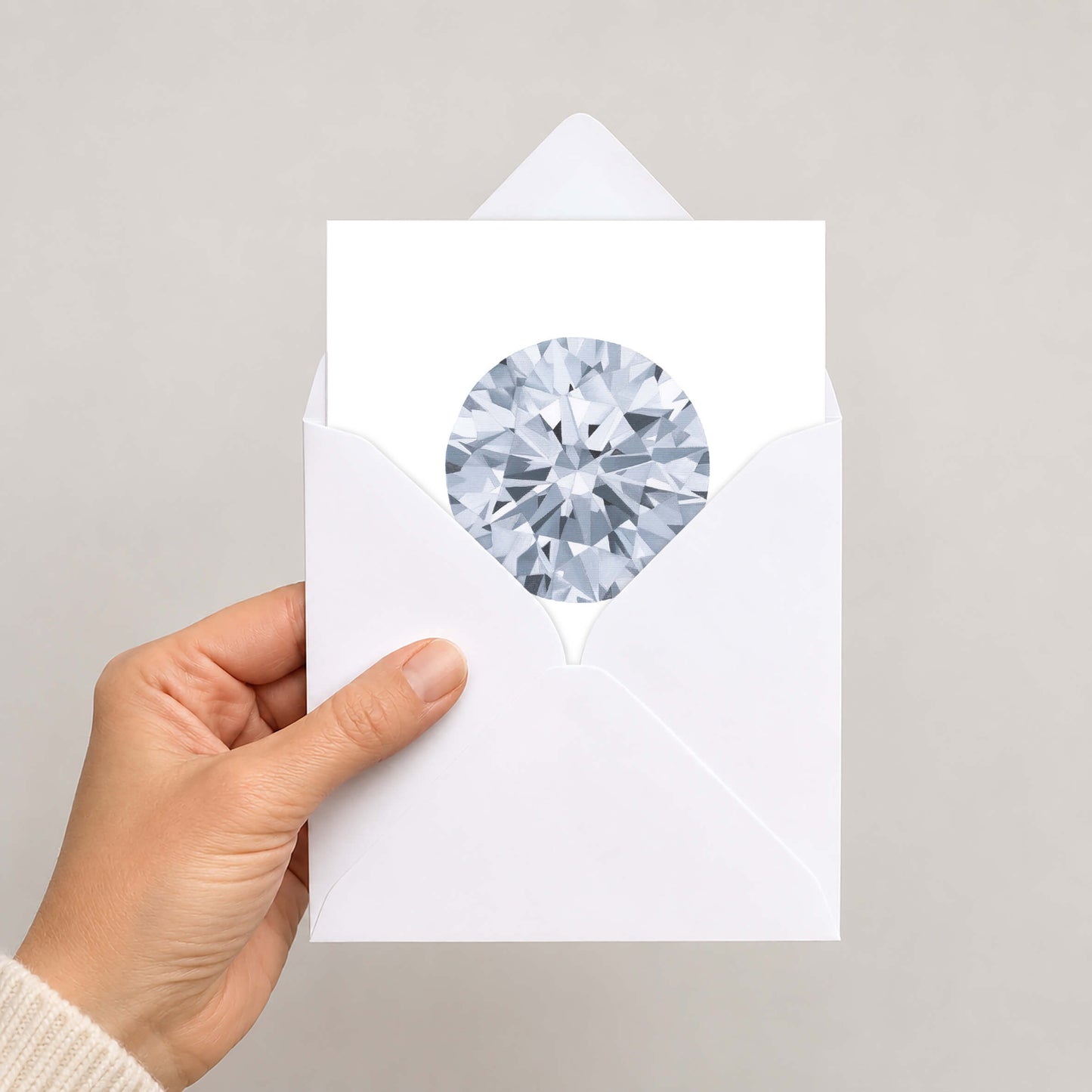 A hand holding a white envelope with a circular cutout showing a detailed, faceted diamond gemstone illustration, set against a plain light grey background.