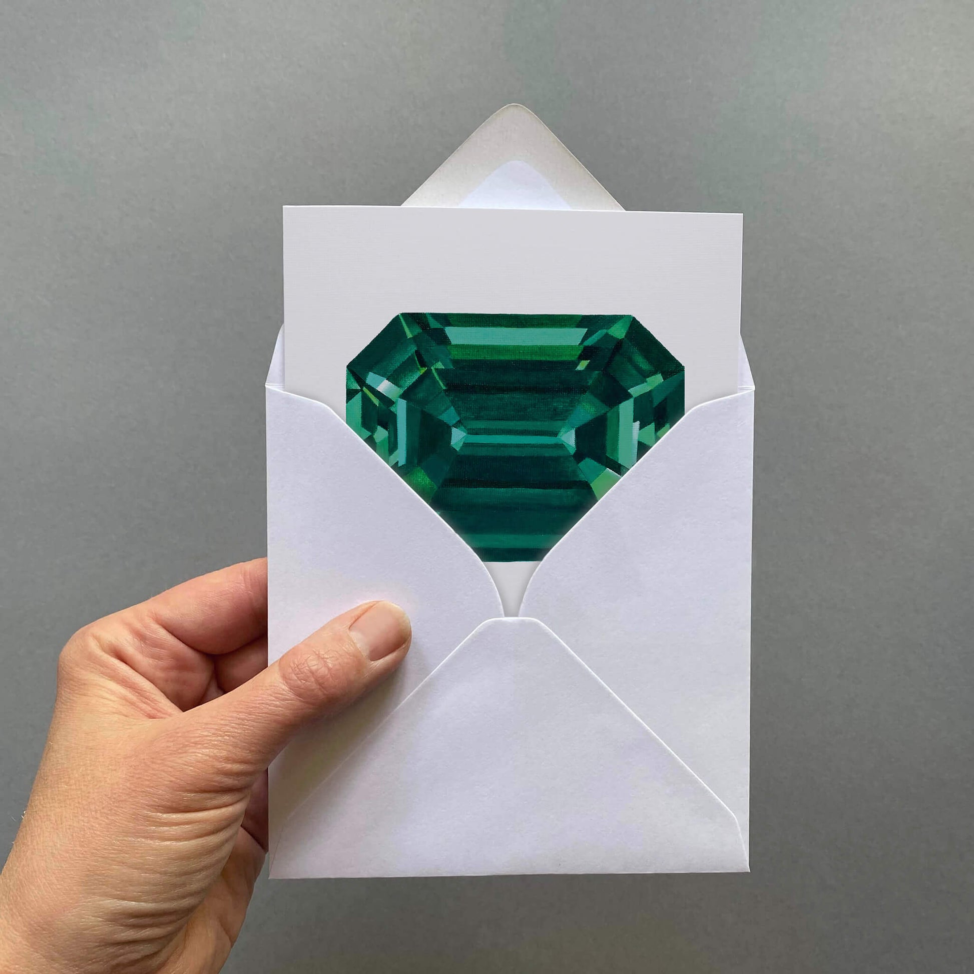 Hand holding a white envelope with a green gemstone card inside against a grey background