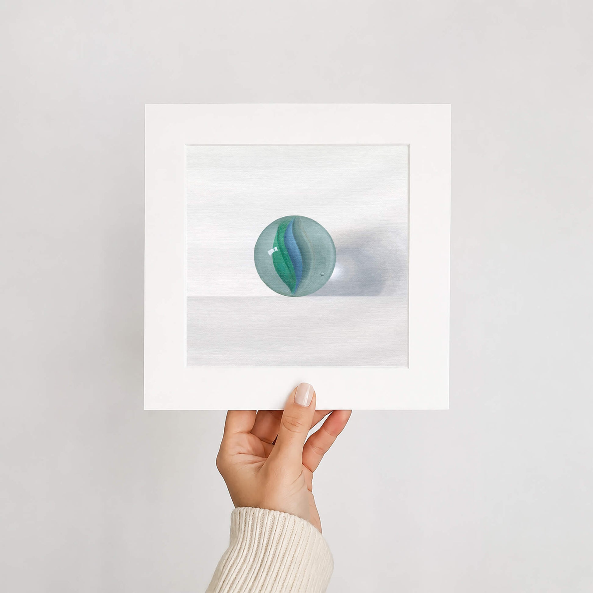 Hand holding a fine art print in mount of glass marble inside against a light gray background