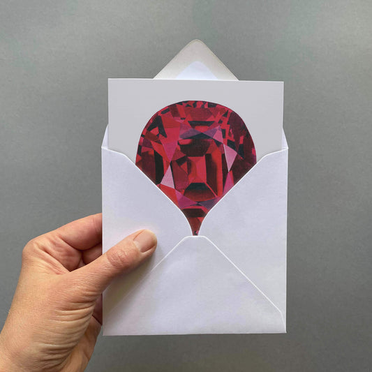 Hand holding a white envelope with a red gemstone design card on a light grey background