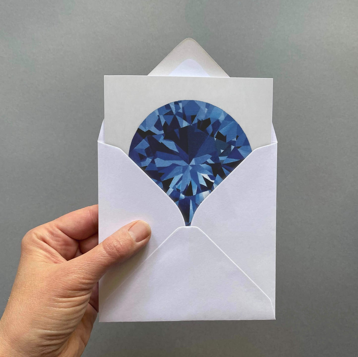 Hand holding a greetings card with a blue sapphire gemstone design on a pale grey background
